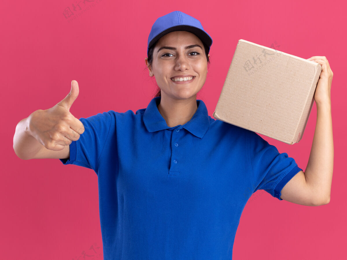 站着面带微笑的年轻送货员穿着制服 肩上扛着帽子盒 在粉红色的墙上孤立地竖起大拇指衣服脸女孩