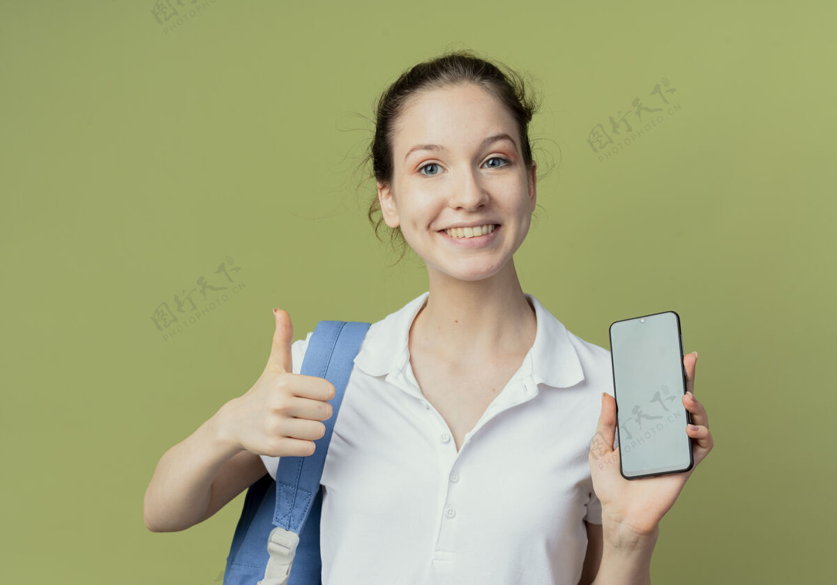 显示带着微笑的年轻漂亮的女学生戴着背袋展示手机和拇指在绿色背景上孤立向上绿色女