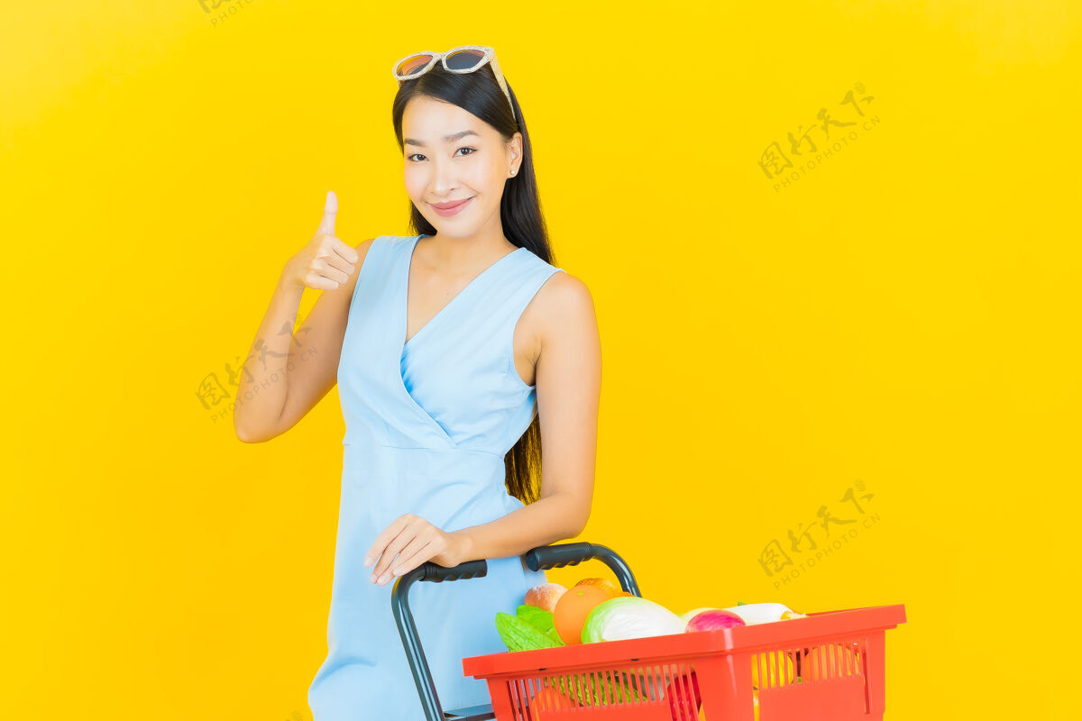 享受一幅美丽的亚洲年轻女子微笑的画像 黄色的墙上挂着超市的购物篮人食物泰国菜