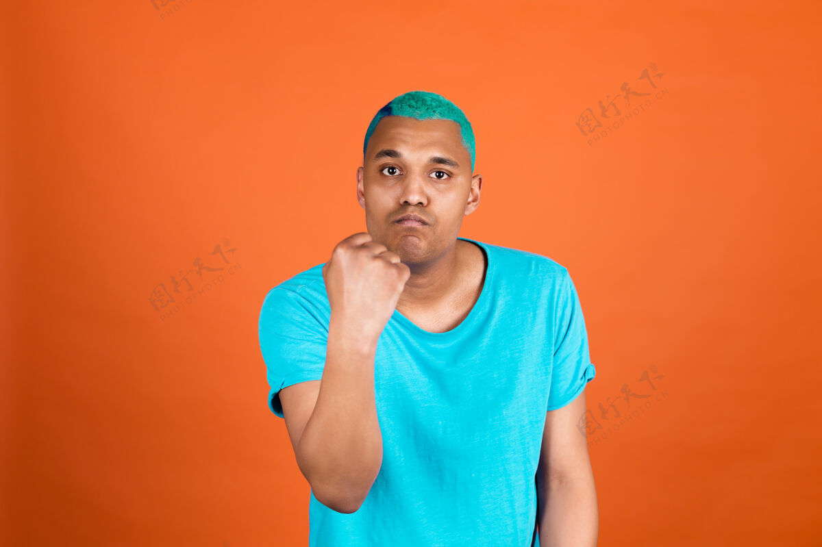 表情黑人非洲男子在随意的橙色墙上蓝色头发大叫愤怒举起拳头不高兴和侵略性工人情绪心烦