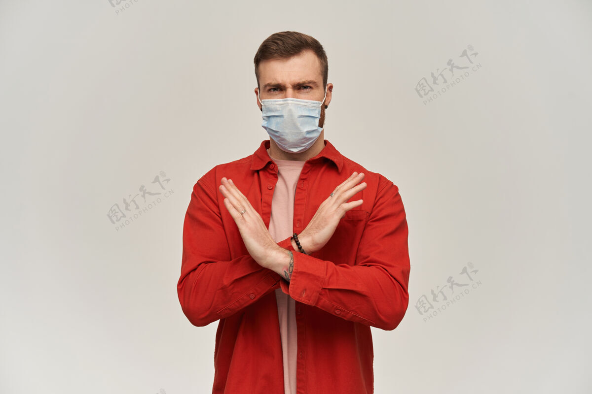 禁止脸上戴着防冠状病毒口罩的严肃的年轻胡须男子用手和胳膊做了一个x形 并在白墙上显示停止标志禁止英俊流行病