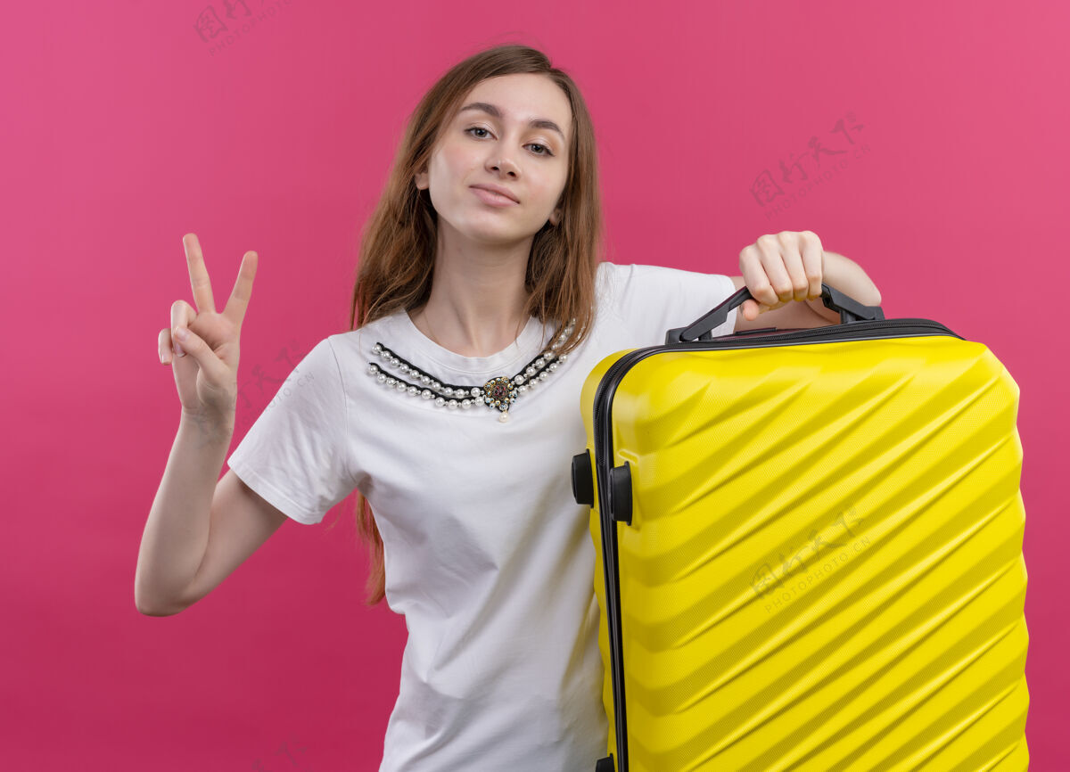 年轻自信的年轻旅行家女孩拿着手提箱在孤立的粉色空间做和平标志标志手提箱信心