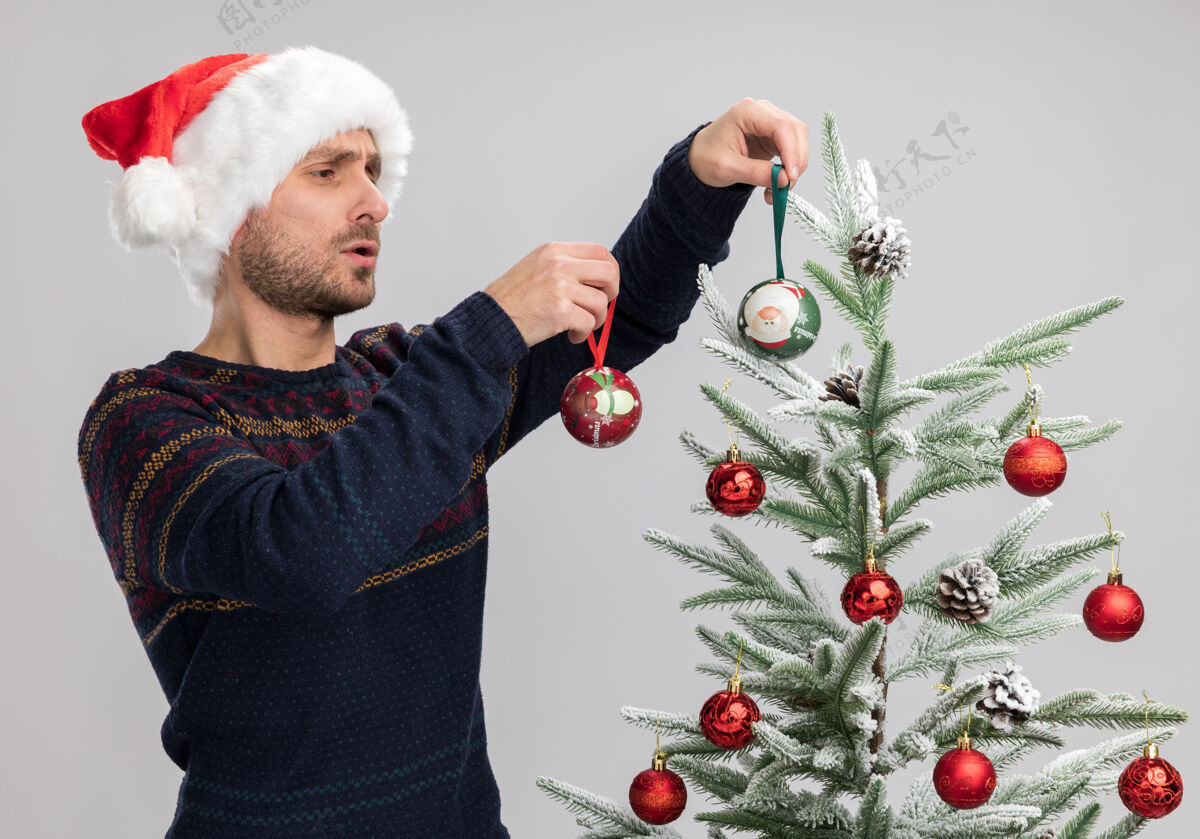装饰品专注的白人年轻人戴着圣诞帽站在圣诞树旁 用白色背景上孤立的圣诞装饰球装饰圣诞树球白色圣诞树