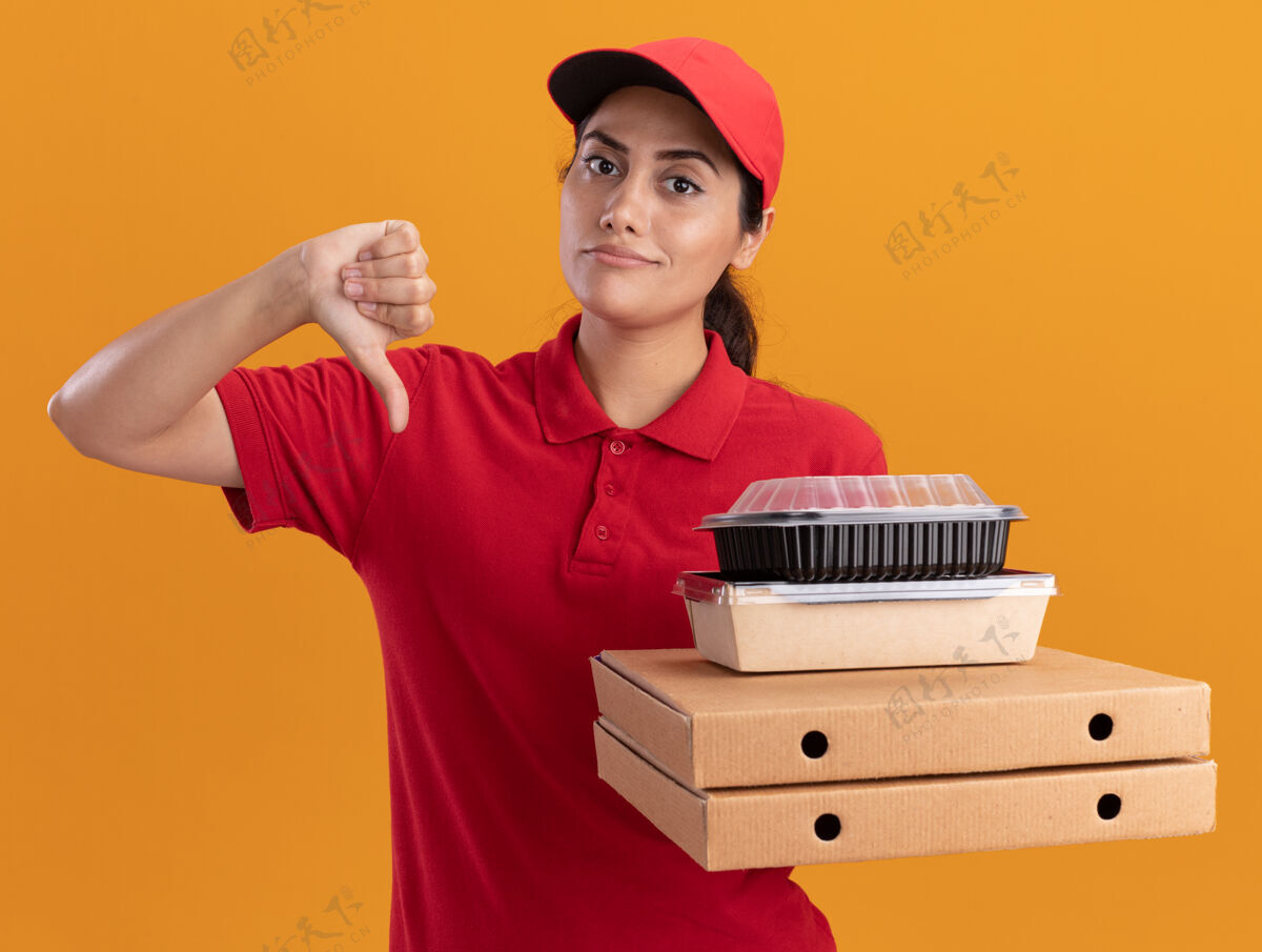 放下令人印象深刻的年轻送货女孩穿着制服 戴着帽子拿着比萨饼盒和食品容器显示拇指向下隔离在橙色墙上表演人感觉