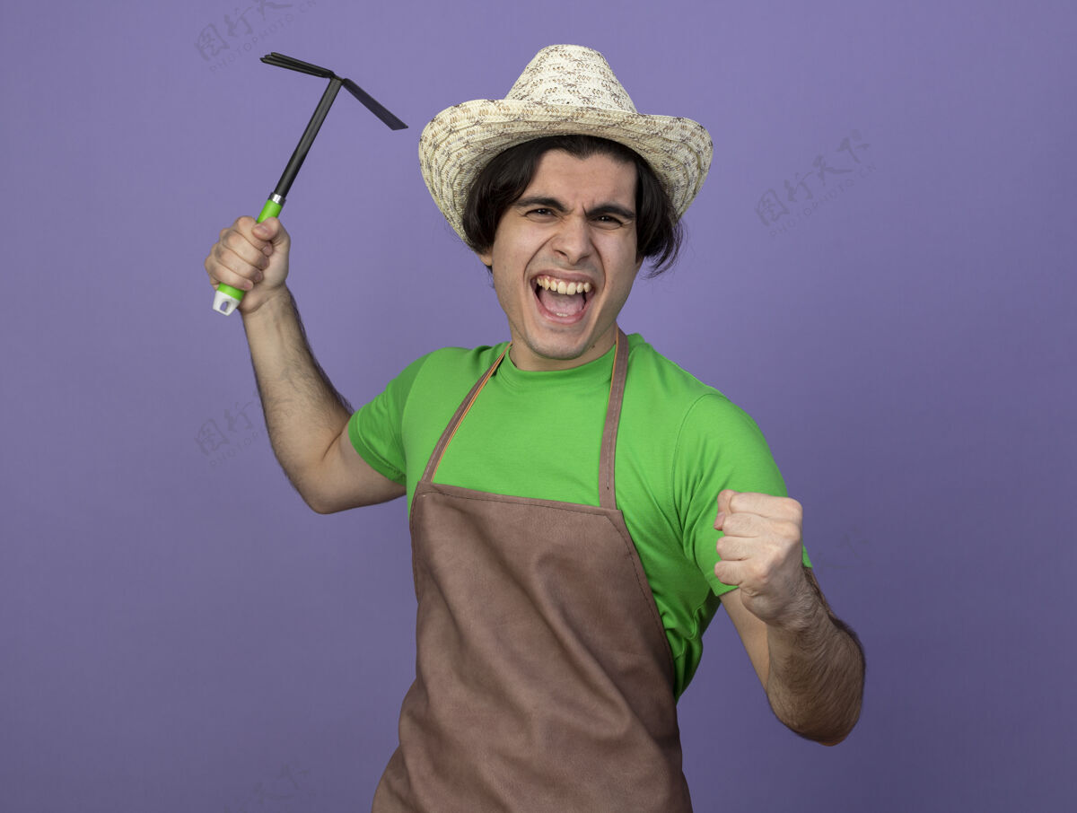手势快乐的年轻男园丁穿着制服戴着园艺帽举起锄头耙表示是的手势园艺男性年轻
