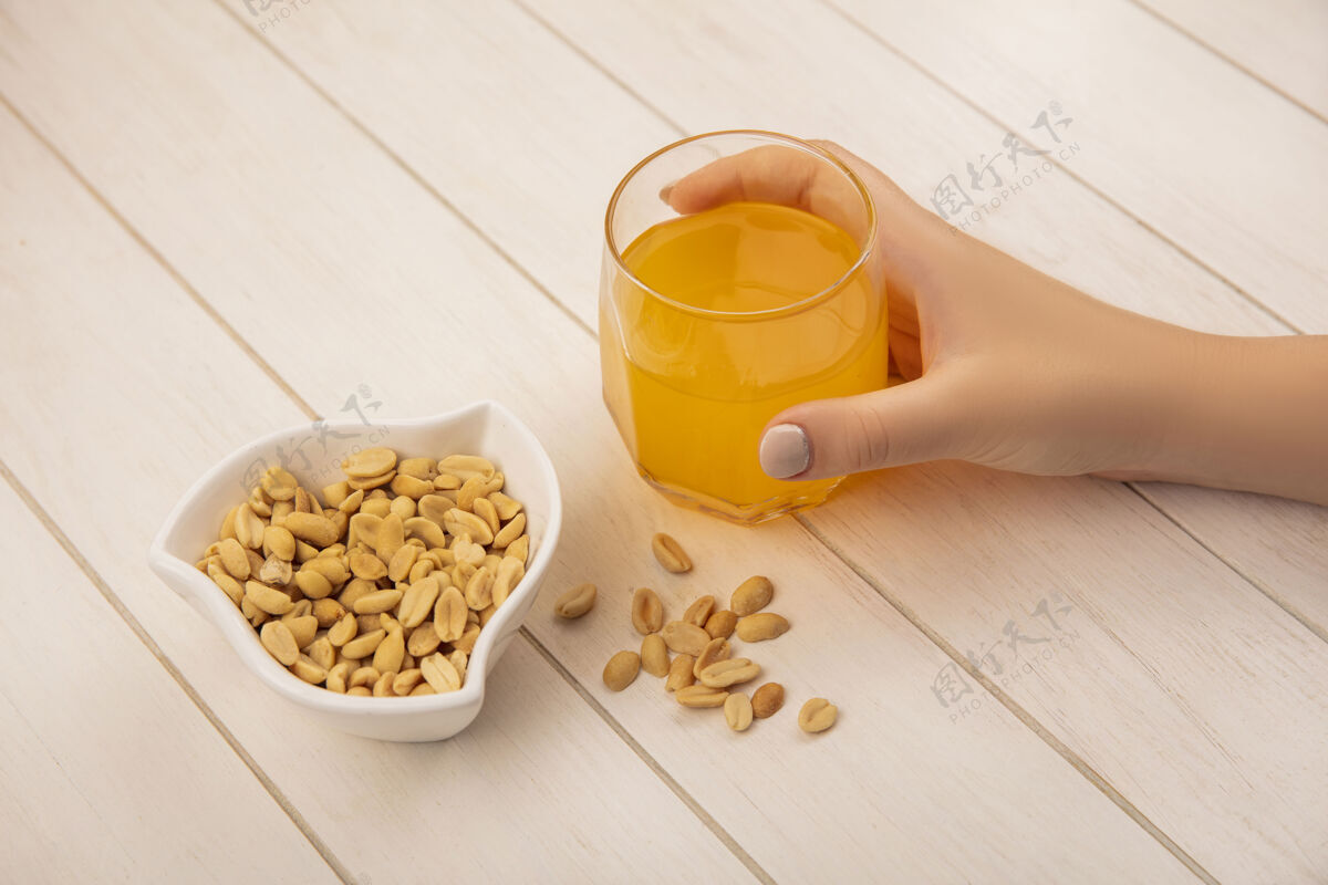 自然女性手拿着一杯橘子汁和松子放在米色木桌上的碗的俯视图米色玻璃视图