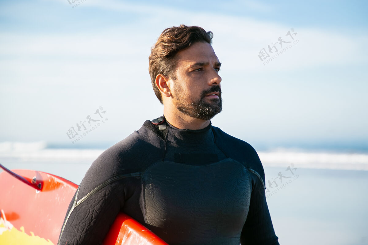 肖像穿着潜水服的白种人运动员拿着冲浪板看着别处站立强壮深色胡子