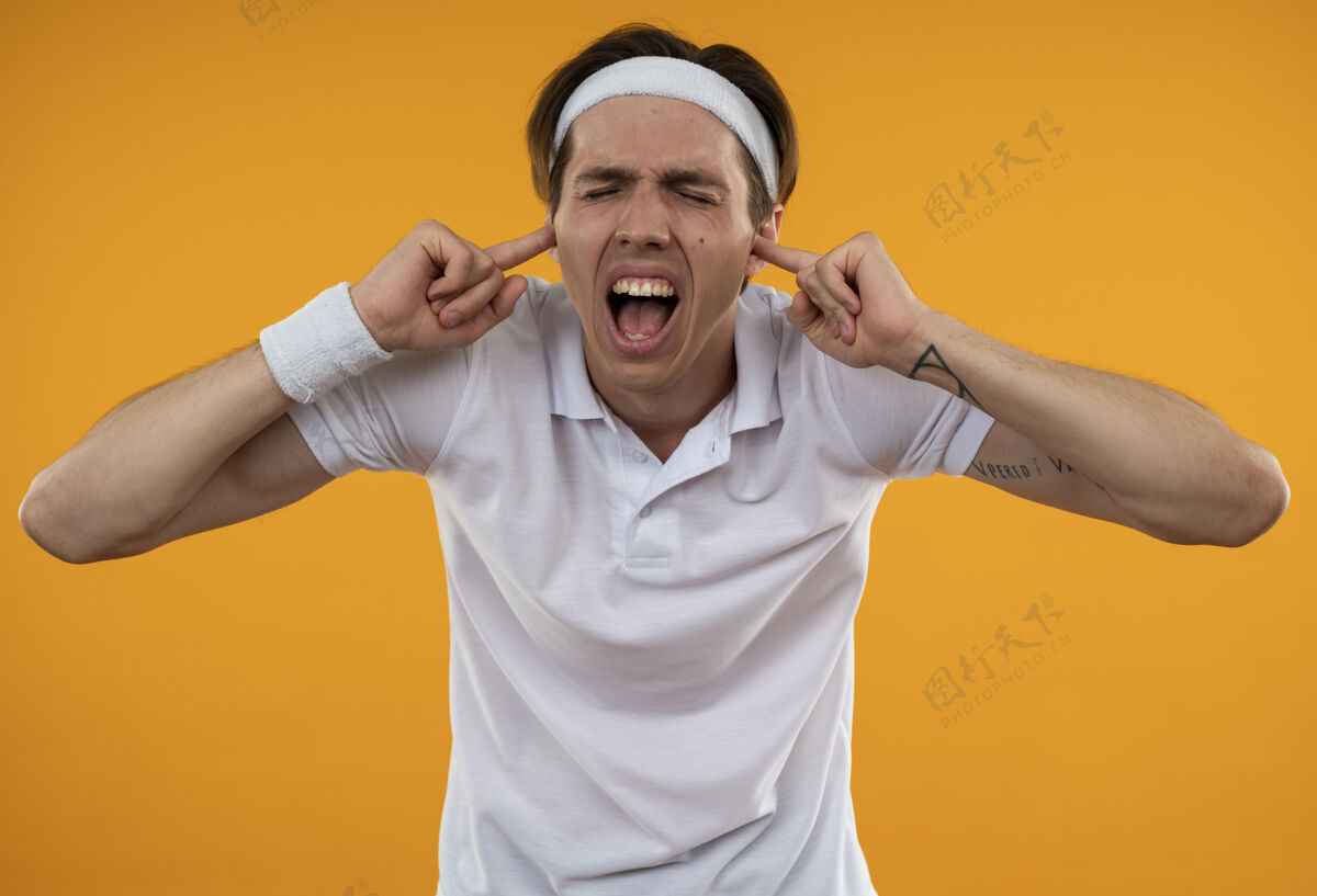 男人愤怒的年轻人闭着眼睛戴着头带和腕带闭着耳朵孤立在橙色的墙上愤怒运动头带