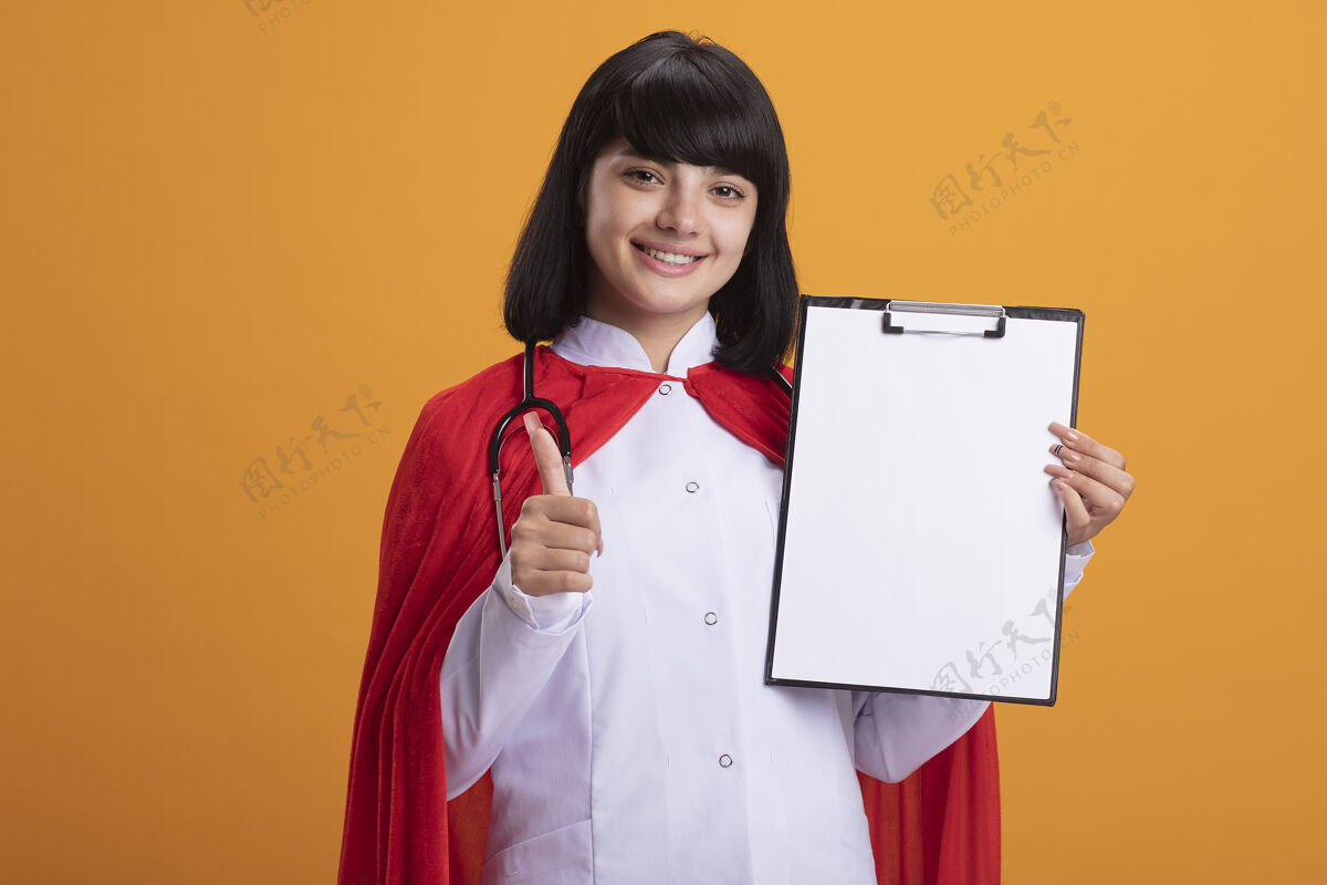 显示带着微笑的年轻超级英雄女孩 穿着听诊器 穿着医用长袍 披着斗篷 拿着剪贴板 在橙色的墙上孤立地竖起大拇指年轻听诊器穿