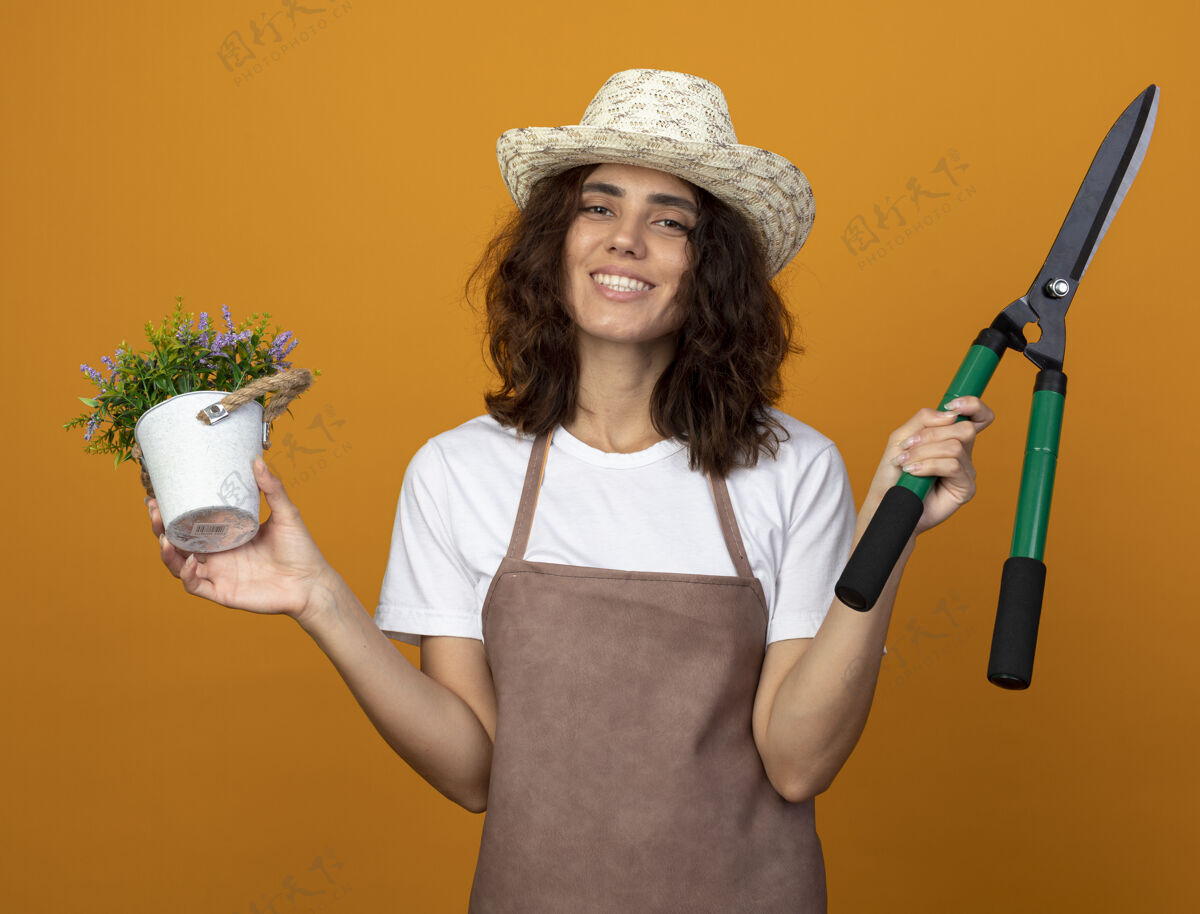 制服身着制服的年轻女园丁微笑着 戴着园艺帽 手里拿着花盆里的花 剪子孤立在橘子上园艺橙剪子