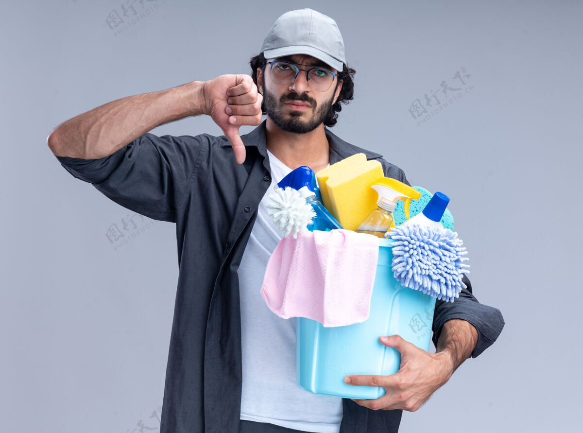 清洁愁眉不展的年轻帅哥 穿着t恤 戴着帽子 手里拿着一桶清洁工具 大拇指朝下 孤零零地躺在白墙上帽子家伙穿