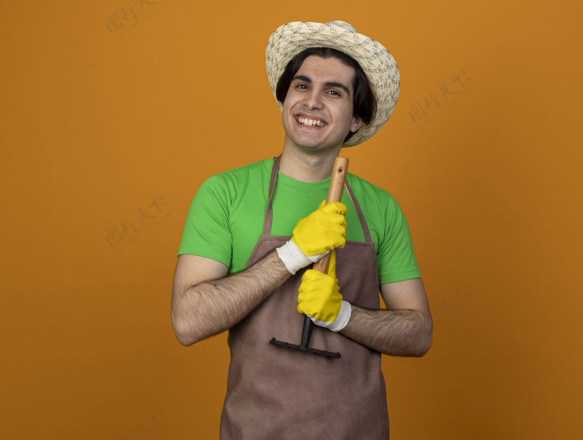 制服年轻的男园丁面带微笑 穿着制服 戴着园艺帽 戴着手套 手里拿着一把橙色的耙子园艺花园年轻