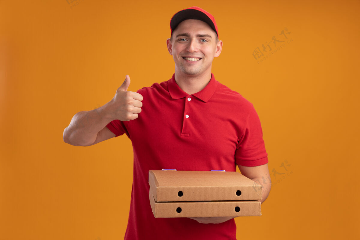 男人微笑的年轻送货员穿着制服 戴着帽子 拿着披萨盒 在橙色的墙上孤立地竖起大拇指盒子穿制服