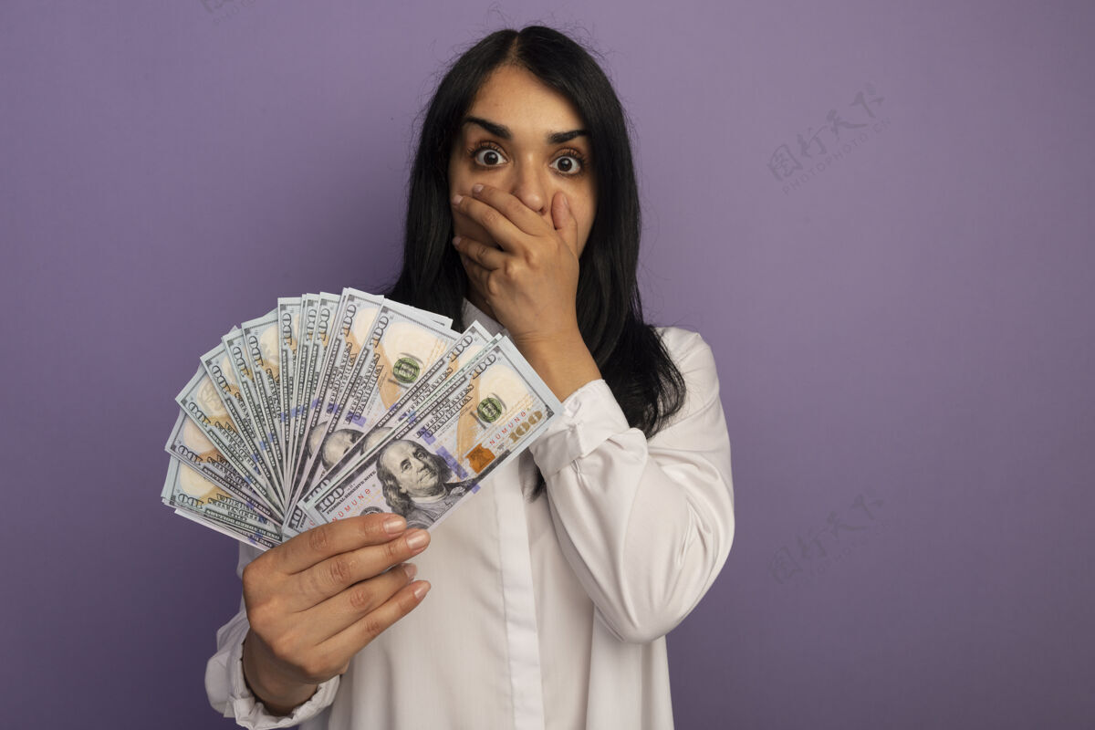 紫色吓得年轻漂亮的女孩穿着白色t恤拿着钱捂着嘴用手隔离在紫色的复印空间上戴着嘴巴年轻
