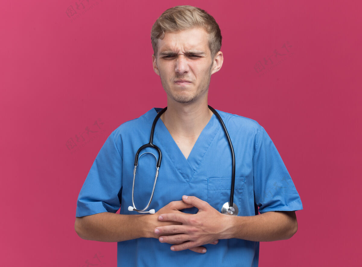 年轻年轻的男医生穿着医生制服 手持听诊器 将手放在疼痛的胃部 隔离在粉红色的墙上制服听诊器手