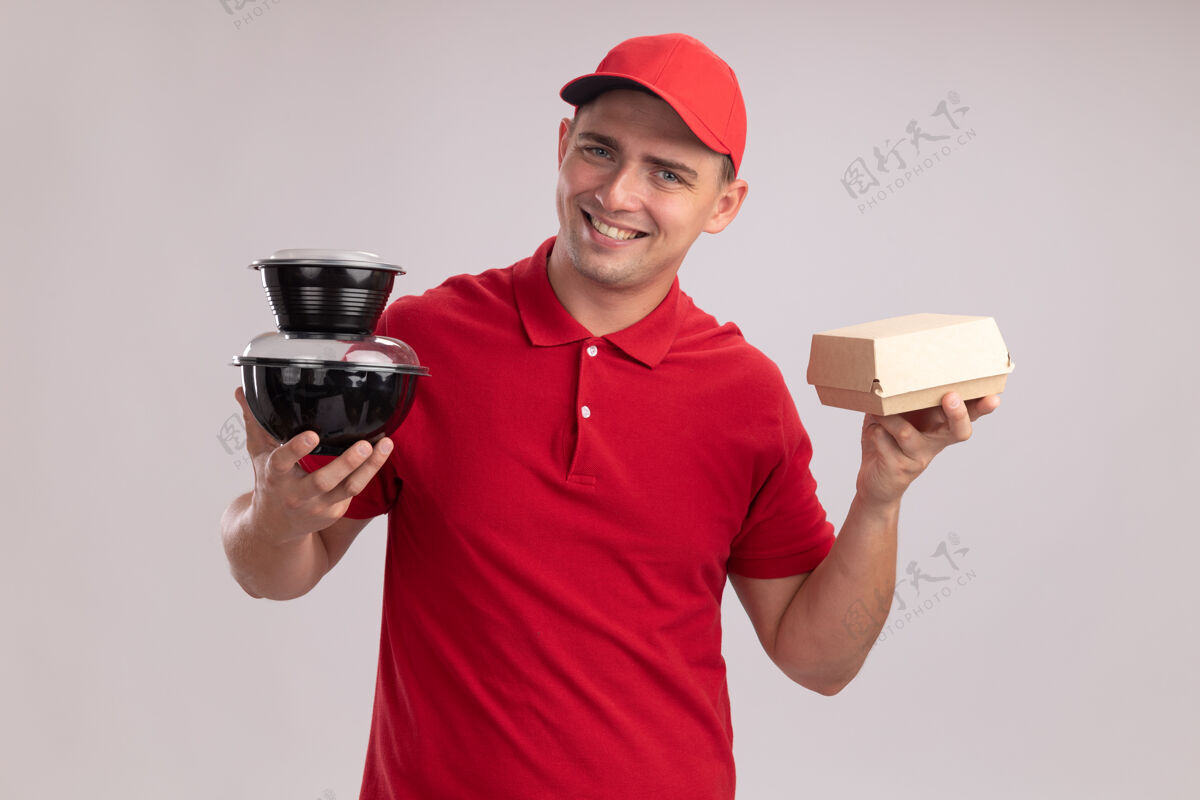 年轻人微笑的年轻送货员穿着制服 戴着帽子 拿着食品容器 纸食品包装隔离在白色的墙上微笑拿着送货