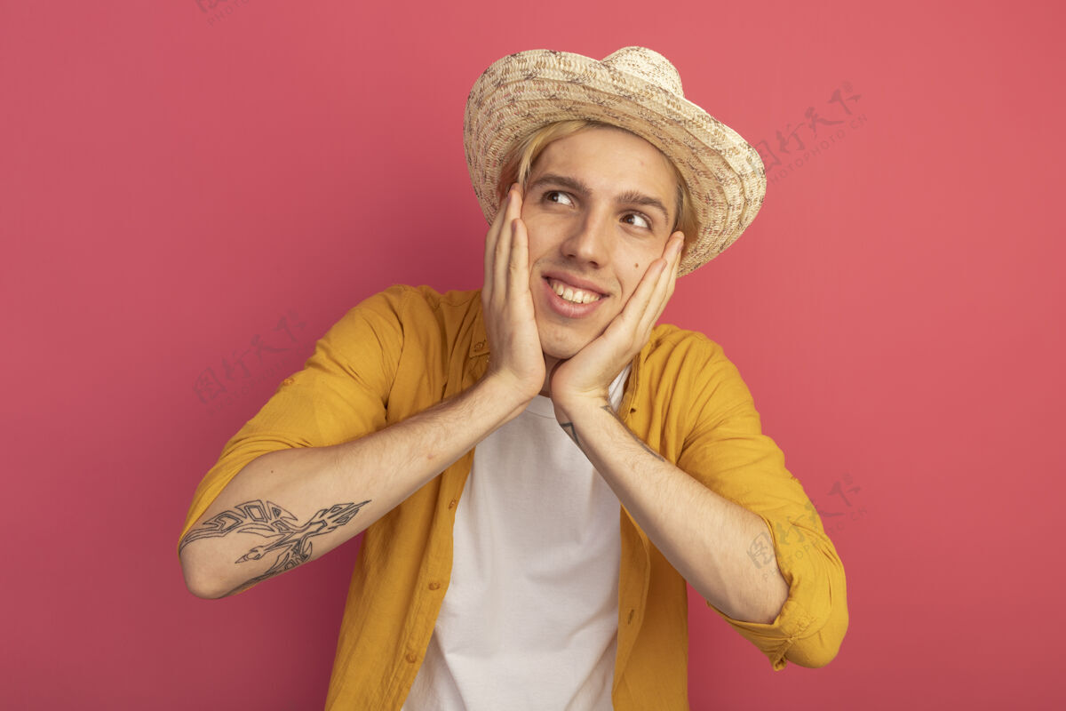 黄色微笑着看着身边年轻的金发小伙穿着黄色t恤 戴着帽子 把手放在脸颊上孤立的粉红色壁板小伙子脸颊