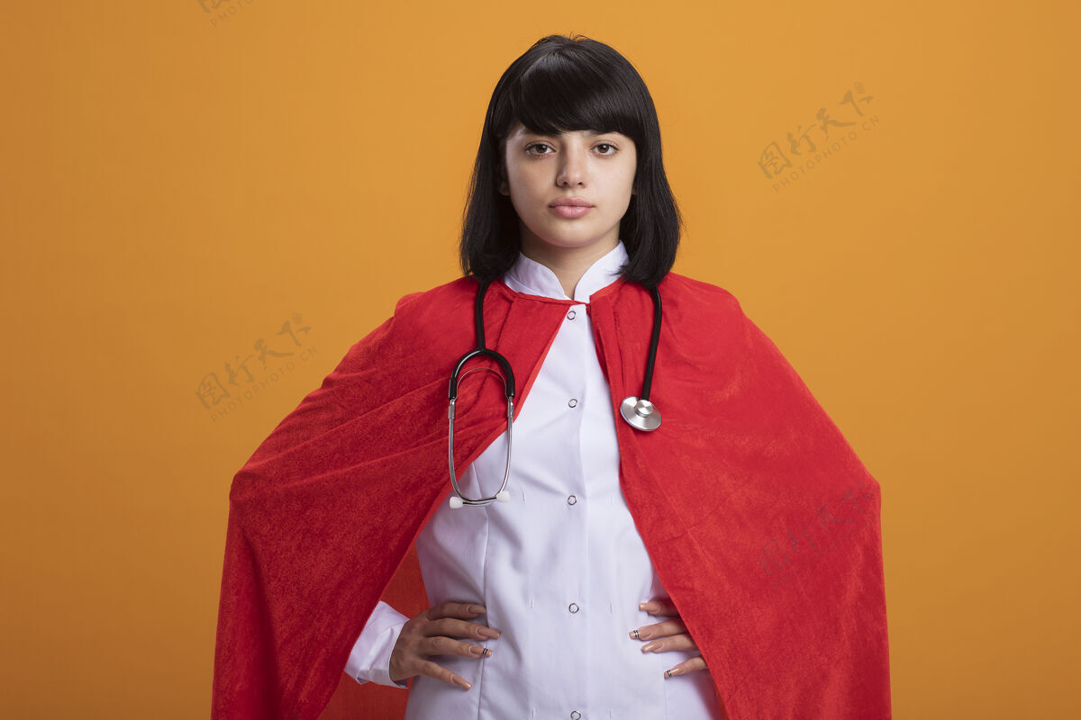 医疗自信的年轻超级英雄女孩戴着听诊器 穿着医用长袍和斗篷 手放在臀部 橙色隔离臀部橙色年轻