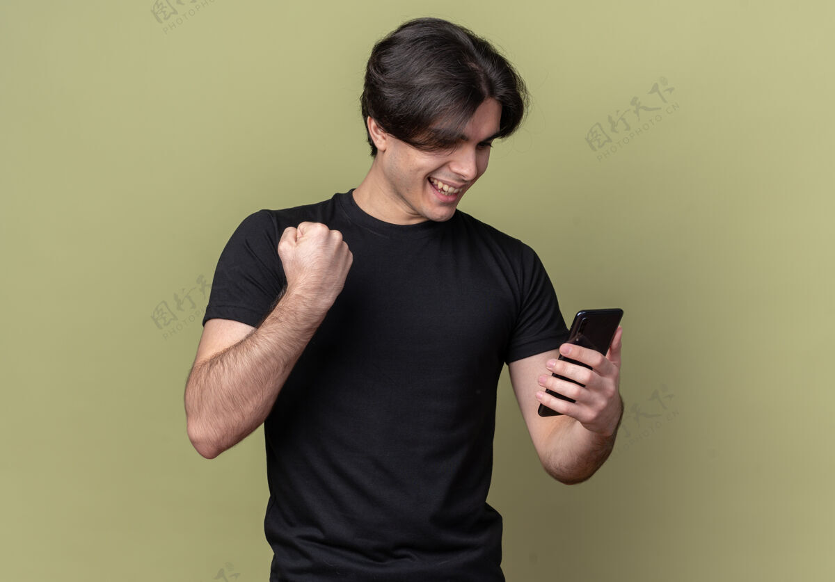 橄榄微笑着的年轻帅哥穿着黑色t恤拿着手机在橄榄绿的墙上显示“是”的手势帅哥年轻穿