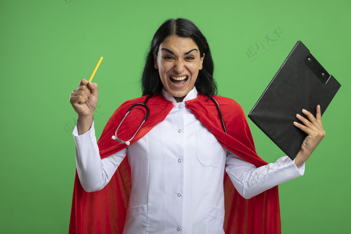 举行兴奋的年轻超级英雄女孩穿着医用长袍 听诊器拿着剪贴板 铅笔隔离在绿色上听诊器长袍兴奋