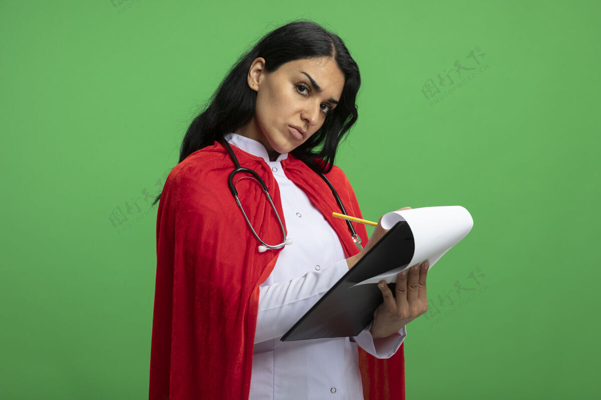 穿严格的年轻超级英雄女孩直视前方 穿着医用长袍 手持听诊器 在绿色的剪贴板上写着什么剪贴板医疗写作