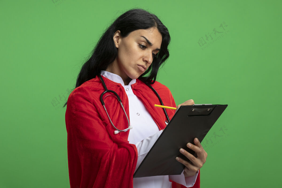 超级英雄严格的年轻超级英雄女孩穿着医用长袍 手持听诊器 在绿色的剪贴板上写着什么严格绿色写作