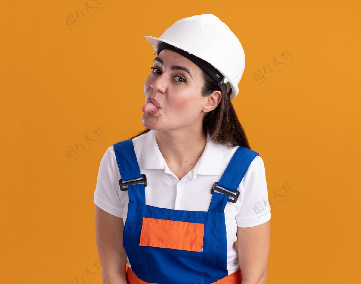 表演穿着制服的年轻建筑工人高兴地在橙色的墙上露出孤立的舌头年轻人拜托舌头