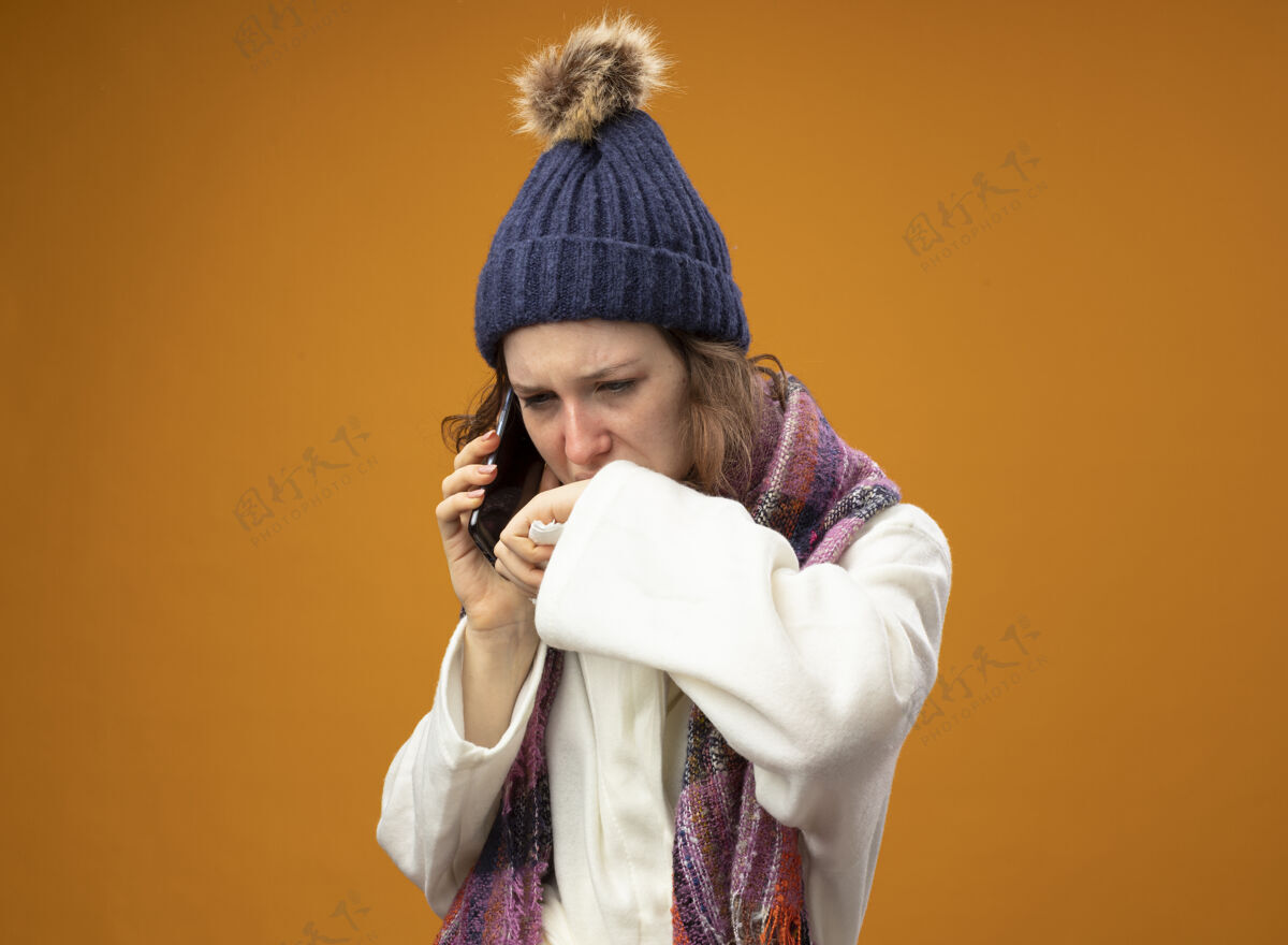 长袍虚弱的年轻生病的女孩看着穿着白色长袍戴着冬天的帽子戴着围巾讲电话擦嘴用手隔离在橙色上橙色嘴巴电话