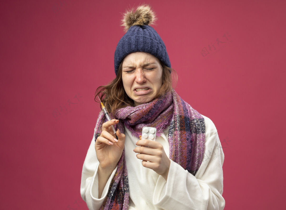 生病哭泣的生病的年轻女孩穿着白色长袍 戴着冬天的帽子 戴着围巾 手里拿着注射器 注射器上隔离着粉红色的药丸年轻药丸帽子