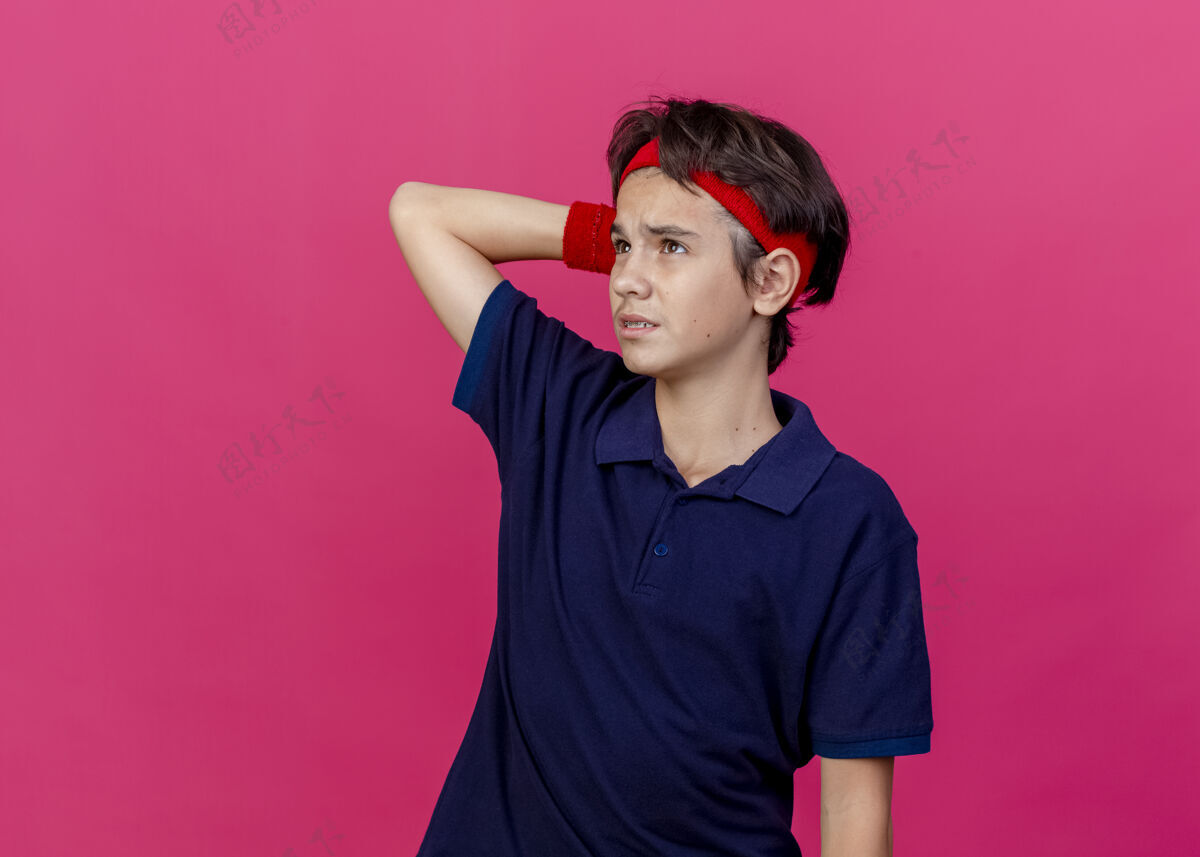 表情体贴的年轻帅气的运动男孩戴着头带和护腕 戴着牙套 抬头仰望 把手放在头后 隔离在粉色的墙上 留着复制空间姿势立场腕带