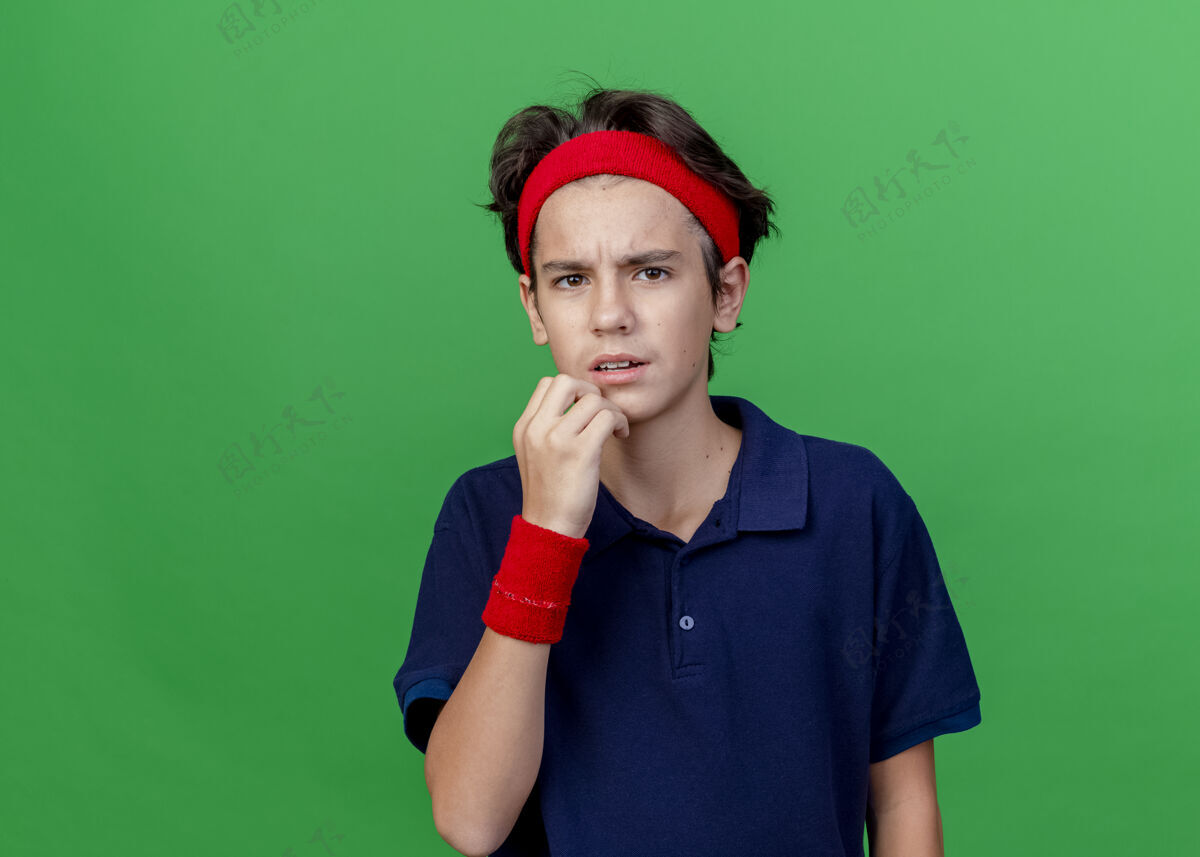 衣服皱眉的年轻帅气的运动男孩戴着头带和护腕 戴着牙套 看着前面 触摸着下巴 隔离在绿色墙壁上 留着复制空间立场腕带男孩