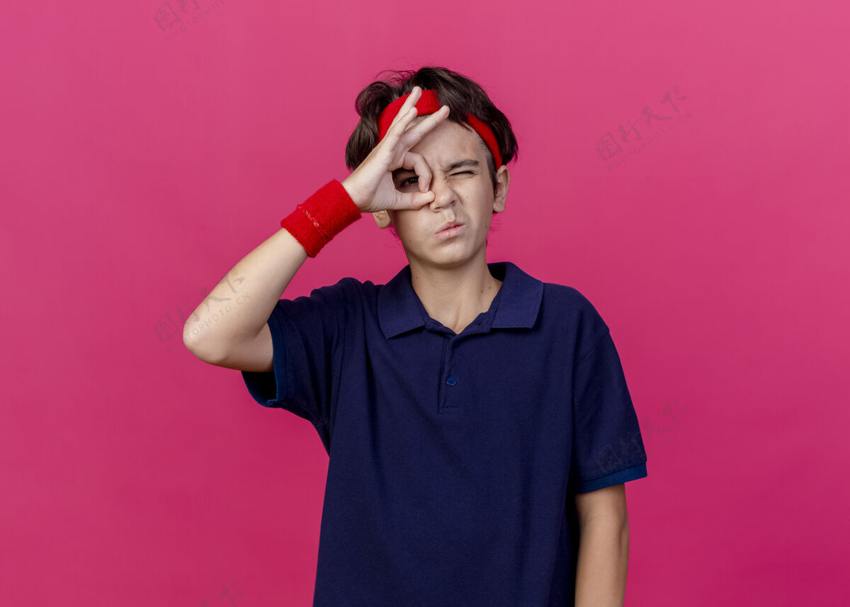 年轻年轻帅气的运动男孩戴着头带和护腕 戴着牙套 在前面眨眼 在粉红色的墙上做着孤立的表情脸市民感觉
