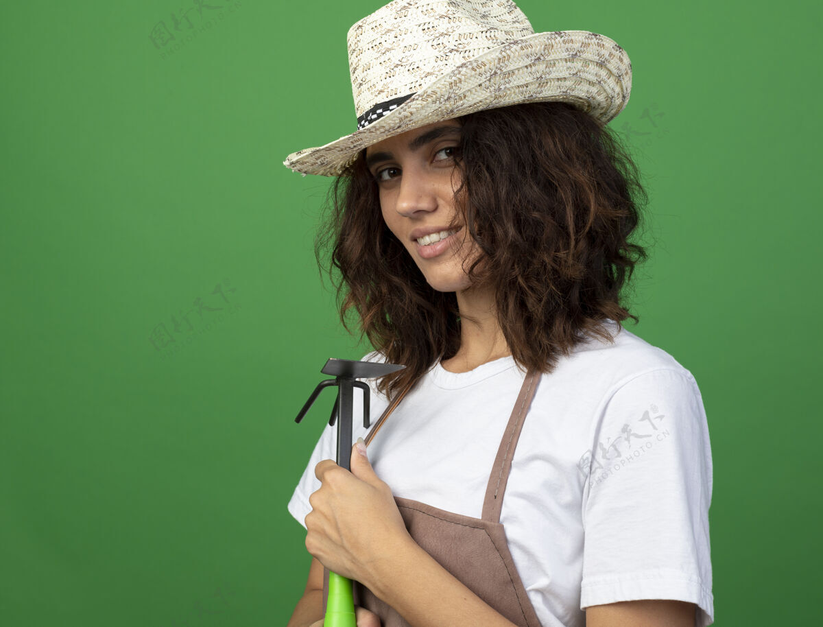 锄头高兴的年轻女园丁穿着制服戴着园艺帽手持锄头耙隔离在绿色耙子穿请