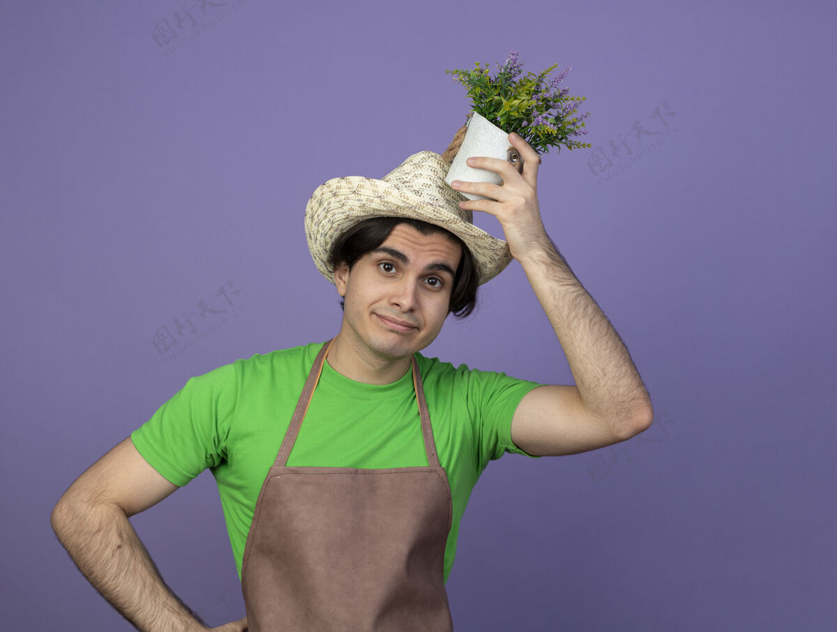 紫色高兴的年轻男园丁穿着制服 戴着园艺帽 头上捧着花盆里的花男人穿花盆