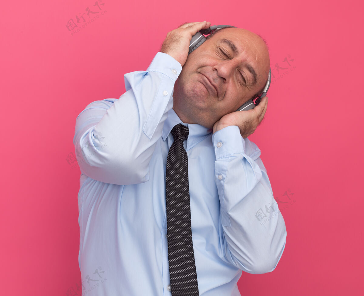 封闭喜欢闭着眼睛歪着头的中年男子 穿着白色t恤 打着领带 把耳机隔离在粉红色的墙上领带中年请