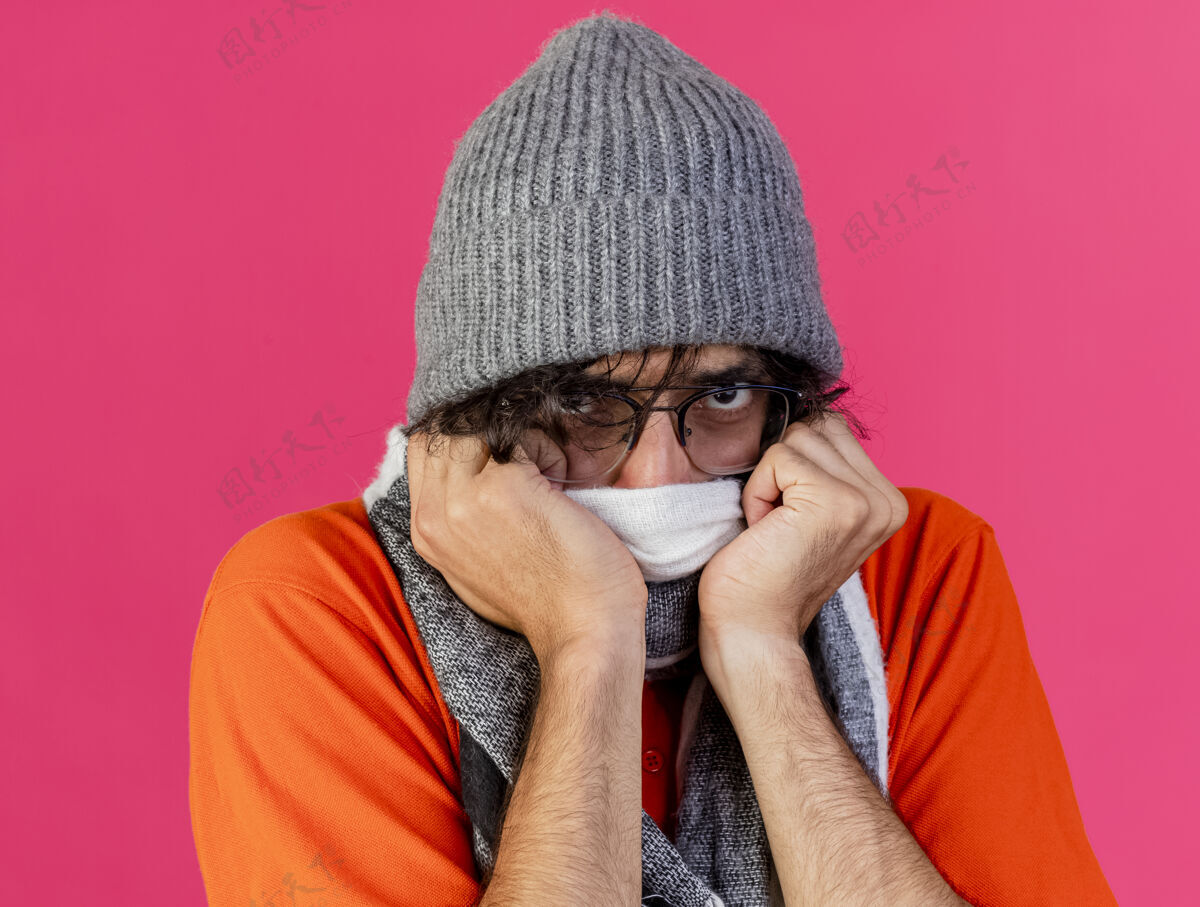 立场特写镜头：愁眉不展的年轻病患戴着眼镜 戴着冬天的帽子和围巾 用围巾捂住嘴 把手放在上面 看着隔离在粉红色墙上的前面表情冬天眼镜