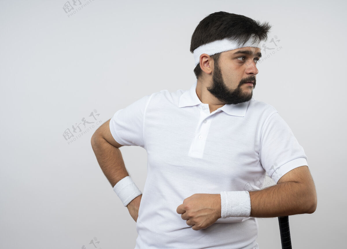 头带年轻的运动型男子一边看一边戴着头带和腕带把手肘放在棒球棒上 隔离在白色墙壁上 留有复制空间肘部棒球穿
