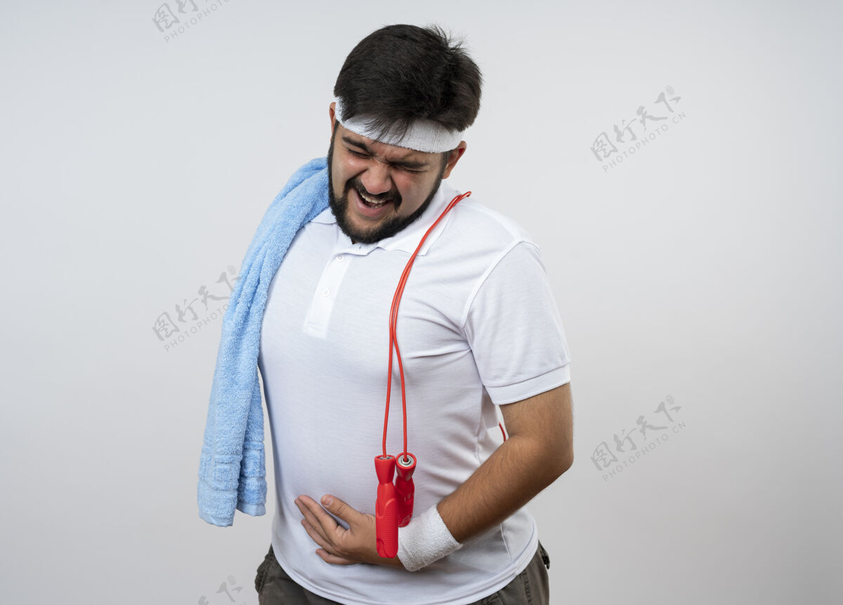 手紧张的年轻人戴着头带和腕带 肩上系着毛巾和跳绳 手放在肚子上 隔离在白墙上男人毛巾紧张