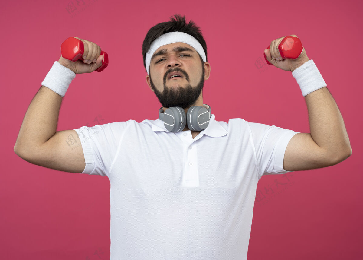 粉红自信的年轻运动型男子戴着头带和腕带 戴着耳机 用粉色的哑铃练习锻炼年轻训练