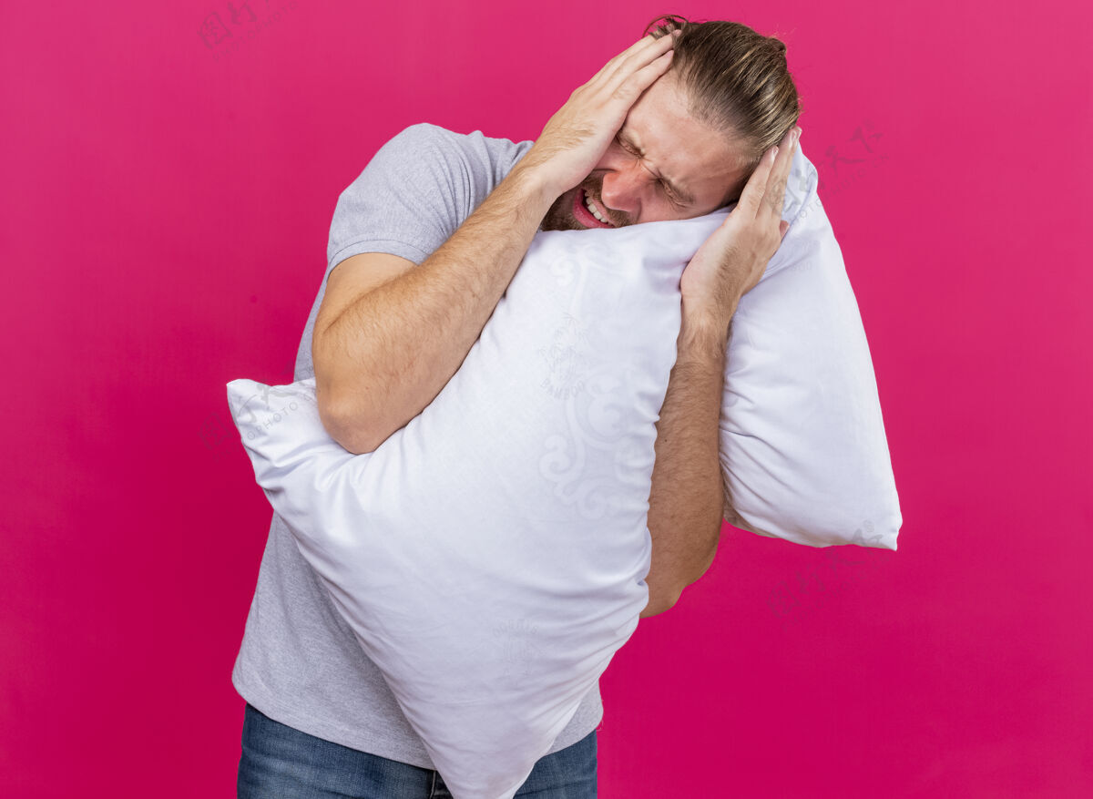 市民疼痛的年轻英俊的斯拉夫病人抱着枕头抱着头闭着眼睛忍受着头痛隔离在粉红色的墙上感觉疾病男人
