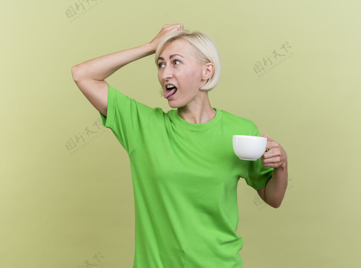 女人恼怒的中年金发斯拉夫女人端着一杯茶 摸着头 看着一边露出的舌头 隔离在橄榄绿的墙上表演杯子金发