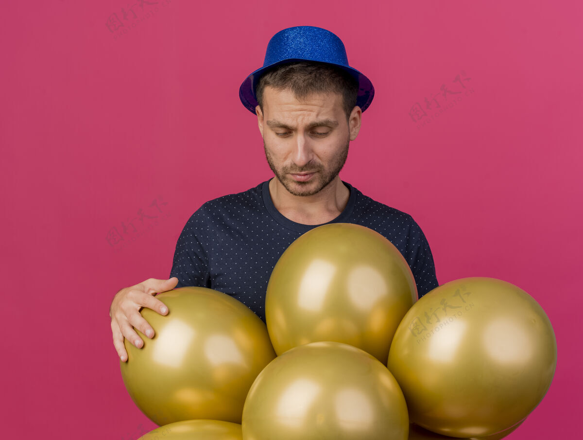 脸戴着蓝色派对帽的帅哥拿着氦气球 隔离在粉红色的墙上 留着复制空间人感觉帅哥