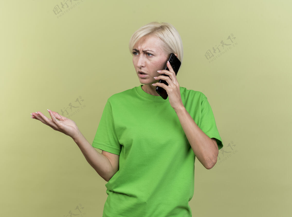 站一位中年金发斯拉夫女人在打电话 一边看一边空手孤立地放在橄榄绿的墙上 留着复印空间金发人不愉快