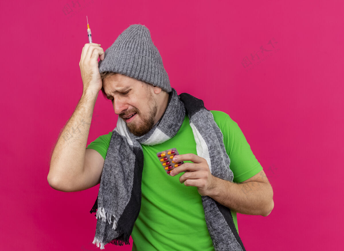 眼睛悲伤而痛苦的年轻英俊的斯拉夫病夫戴着冬天的帽子和围巾拿着注射器和一包胶囊摸着头闭着眼睛隔离在粉红色的墙上有复印空间人斯拉夫人人