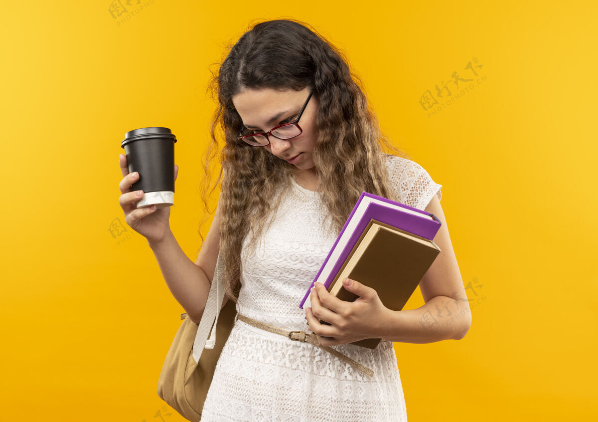 咖啡年轻漂亮的女学生戴着眼镜 背着袋子 手里拿着塑料咖啡杯和书 在黄色的背景上 看着孤立的复印空间塑料杯子漂亮