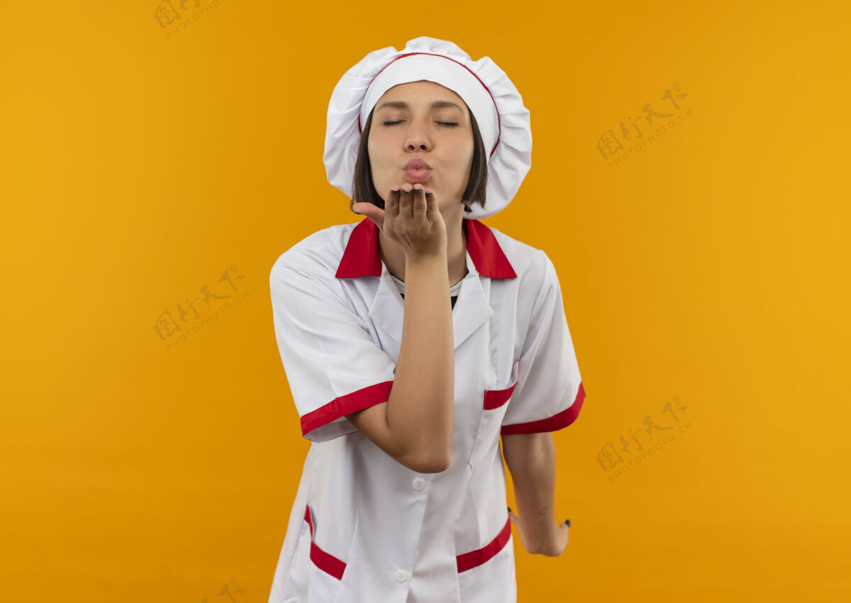 年轻身着厨师制服的年轻女厨师在橙色背景上闭着眼睛对着镜头吹吻发送烹饪眼睛
