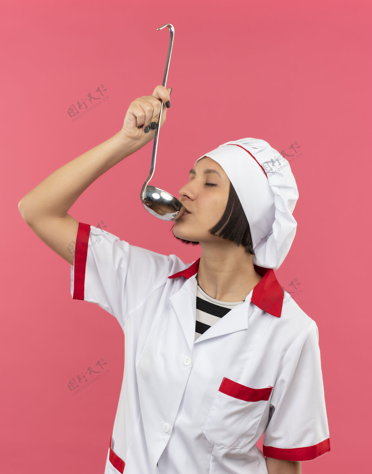 封闭身着厨师制服的年轻女厨师从勺子里喝水 闭着眼睛 在粉色背景上与世隔绝年轻饮用厨师