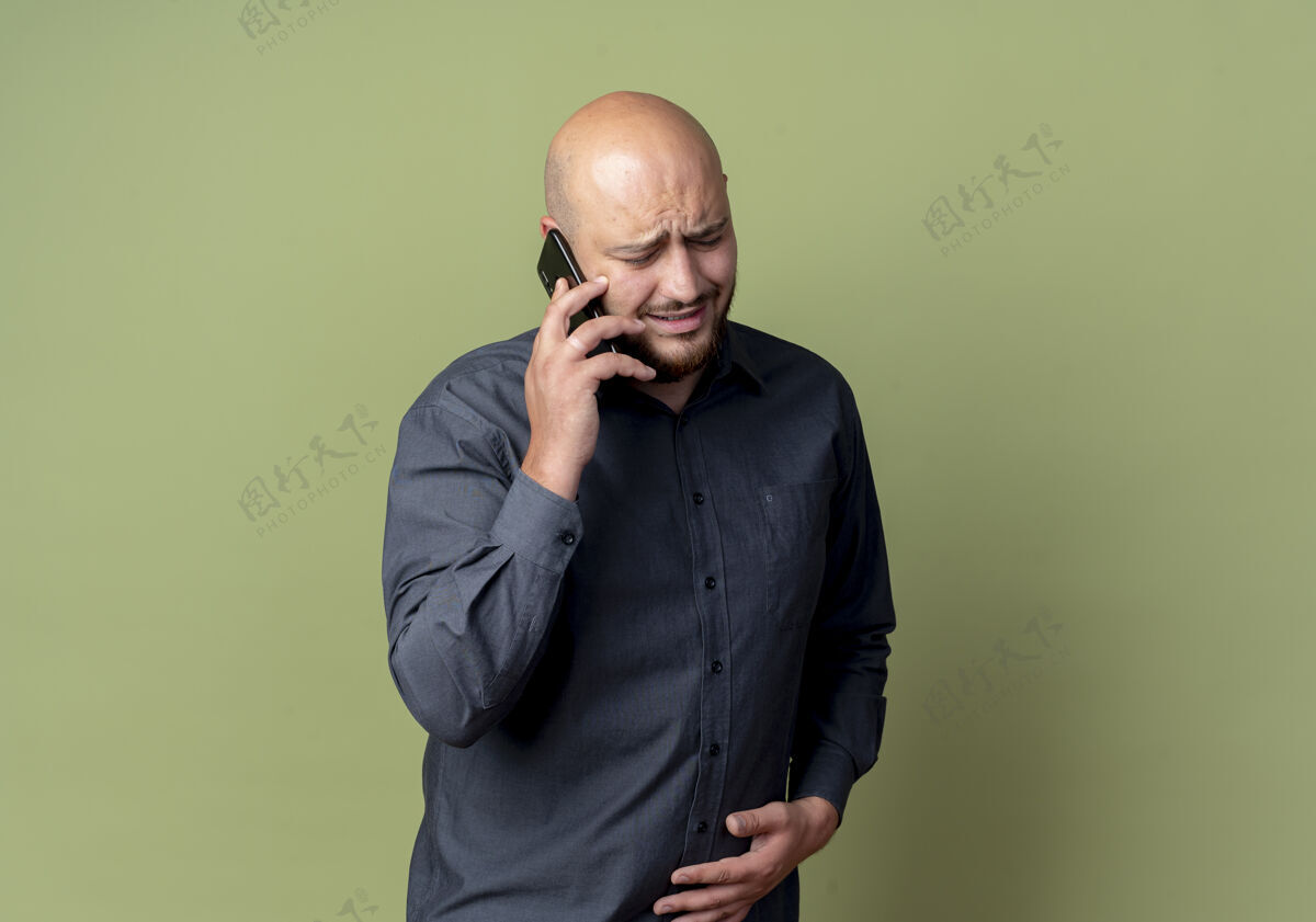 中心未出租的年轻秃顶呼叫中心男子在电话上说话 手放在肚子上孤立的橄榄绿背景与复制空间不愉快年轻电话