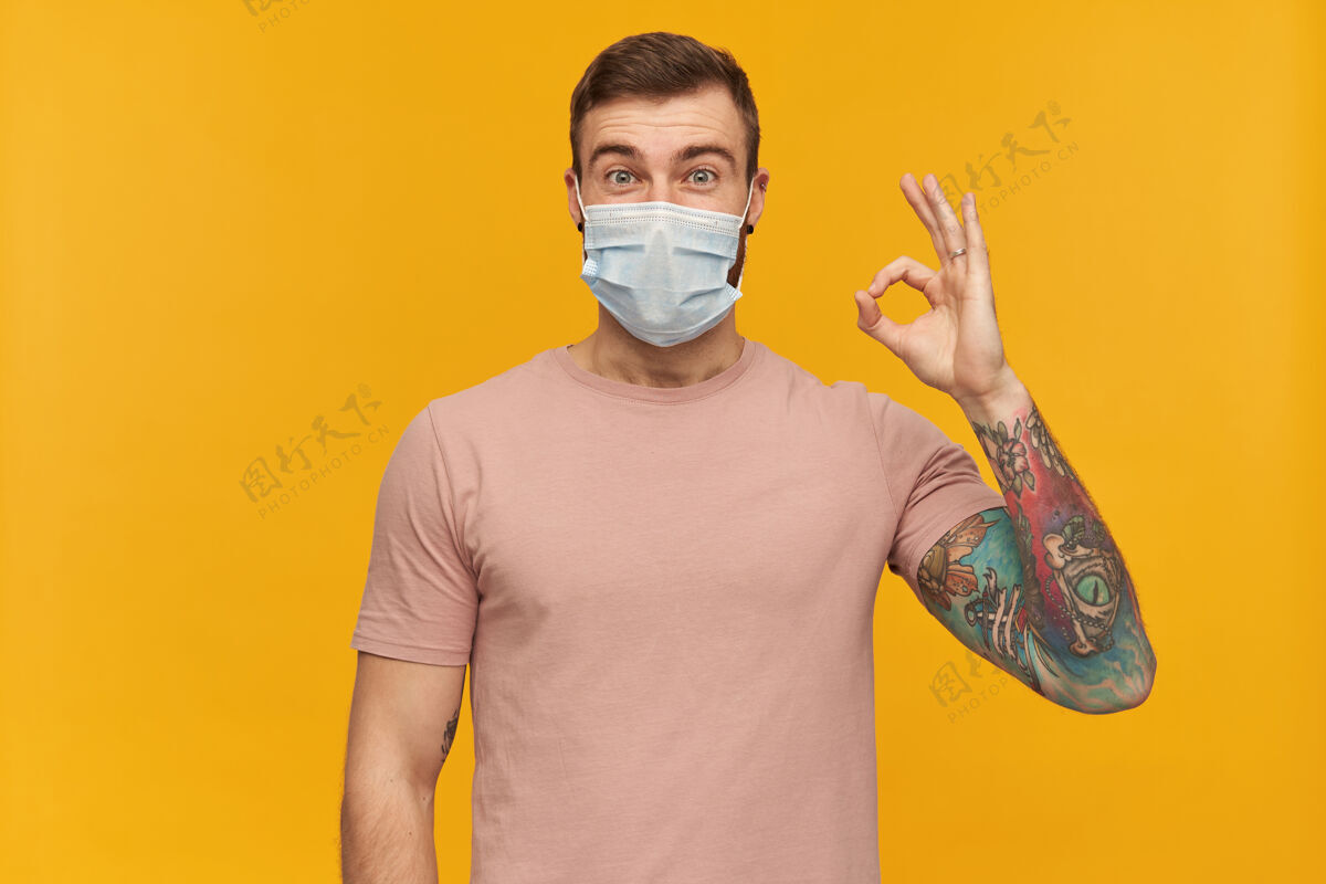 流行病兴奋的纹身年轻人穿着粉色T恤 脸上戴着防病毒面具 脸上留着防冠状病毒的胡子 站在黄色的墙上 脸上有ok标志男性兴奋可以
