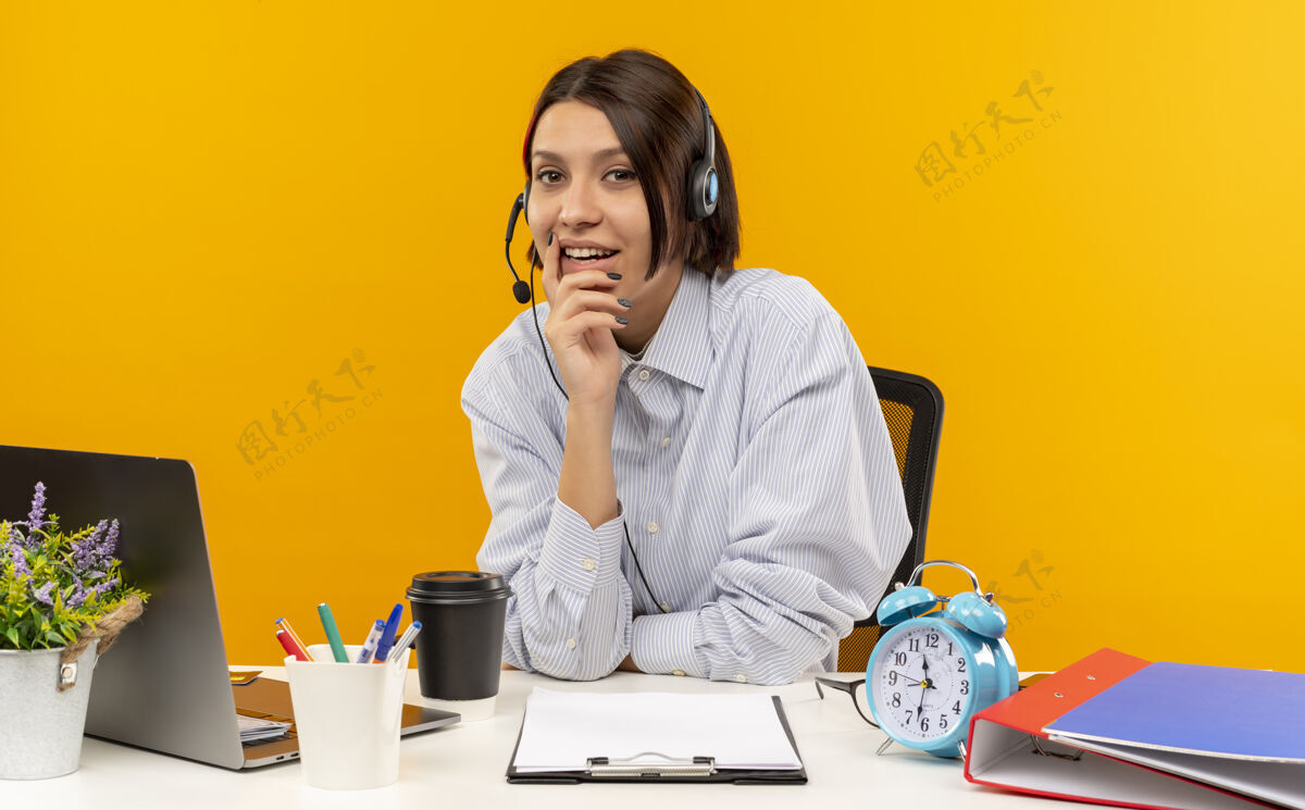 请戴上高兴的年轻呼叫中心女孩戴着耳机坐在办公桌旁把手放在下巴上孤立的橙色背景耳机手女孩